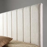 An upholstered headboard in a textured off-white fabric with vertical channel tufting, shown with a bed and a brown pillow in a neutral gray room setting.
