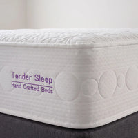 A close-up of a white mattress corner with quilted circular and leaf-like stitching, showing the text 'Tender Sleep Hand Crafted Beds' in purple on the side. The mattress sits on a dark gray bed base against a light neutral background.
