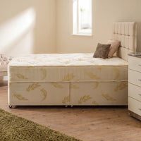 A single bed with a tufted cream headboard and upholstered base, shown in a softly lit bedroom. The mattress and base are cream with a subtle gold leaf pattern, styled with two pillows.