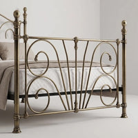 An ornate metal bed frame in a warm antique brass finish, shown with white bedding in a minimalist bedroom setting.