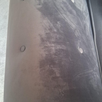 A close-up of a dark gray upholstered surface with visible wear, scuffs, and a small circular hole, photographed against a light gray floor/background.