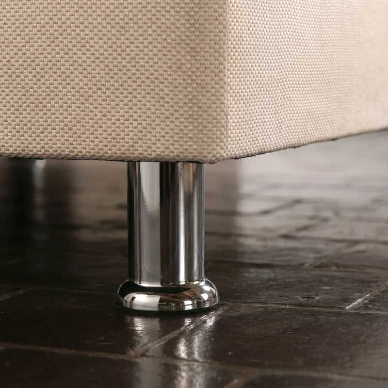 Close-up of a beige woven fabric upholstered furniture corner resting on a polished chrome metal leg over a dark tiled floor.