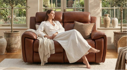 Woman in a white outfit sitting on a brown leather sofa in a bright living room.