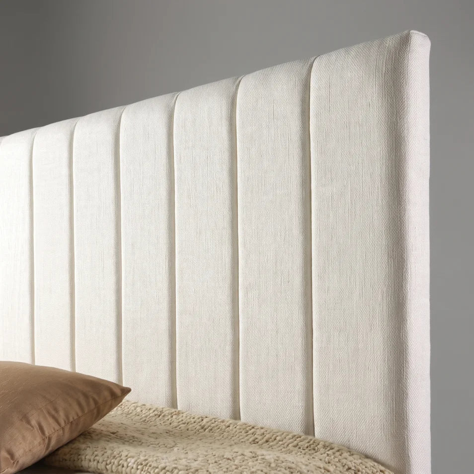 An upholstered headboard in a textured off-white fabric with vertical channel tufting, shown with a bed and a brown pillow in a neutral gray room setting.