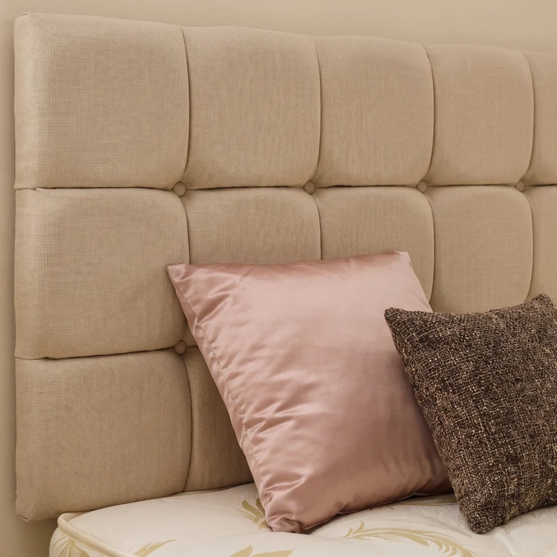 Beige tufted upholstered headboard with two decorative pillows, one dusty pink satin pillow and one dark brown textured pillow, set against a light beige wall.
