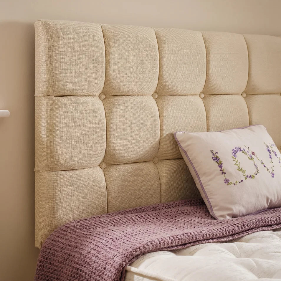 A large tufted upholstered headboard in a warm cream-beige fabric, shown on a bed with a mauve knit throw blanket and a decorative pillow.