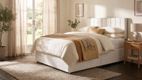 A white upholstered bed frame with a tall segmented headboard, dressed in cream bedding and a tan throw blanket, shown in a softly lit bedroom with curtains, a nightstand, and a rug.