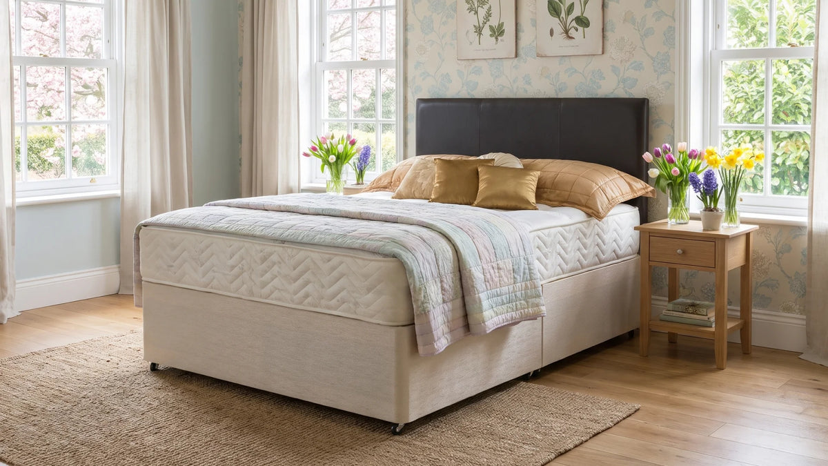 A bed with a cream upholstered base and a dark headboard, dressed in beige and white bedding, set in a bright bedroom with windows, wallpaper, a side table, and flowers.