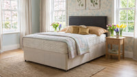 A bed with a cream upholstered base and a dark headboard, dressed in beige and white bedding, set in a bright bedroom with windows, wallpaper, a side table, and flowers.
