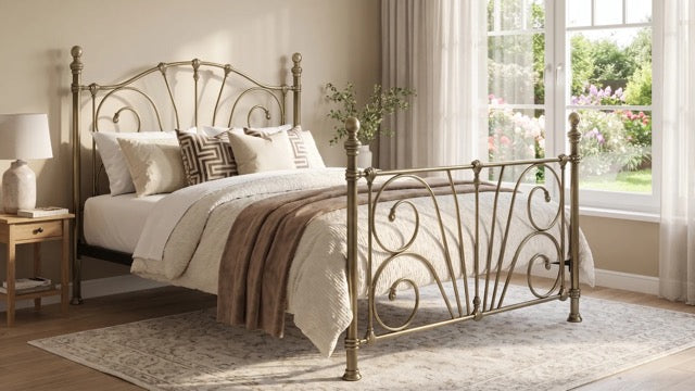 An ornate metal bed frame in a warm champagne-gold finish, styled in a bright bedroom with beige bedding, patterned pillows, a brown throw blanket, a wooden nightstand with a lamp, and a large window looking out to a garden.