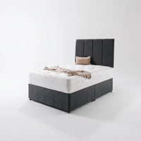 A dark gray upholstered divan bed frame with a tall segmented headboard, shown with a white mattress, a beige pillow, and a beige throw blanket on a clean white studio background.