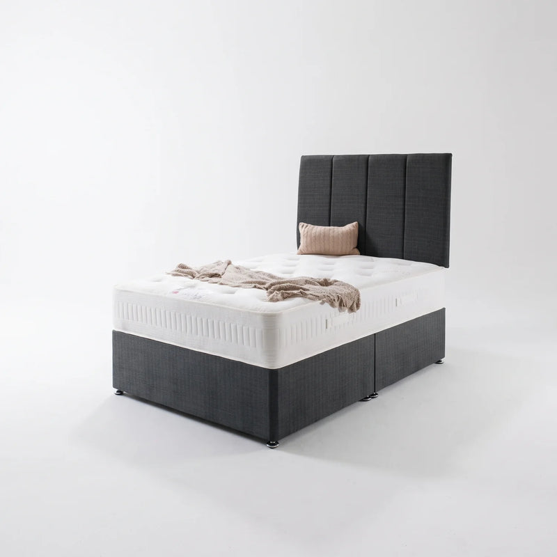 A dark gray upholstered divan bed frame with a tall segmented headboard, shown with a white mattress, a beige pillow, and a beige throw blanket on a clean white studio background.