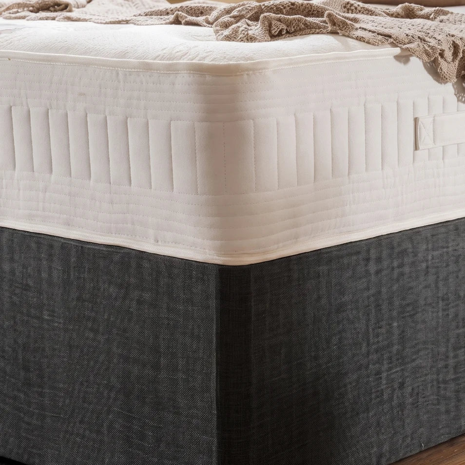 A close-up of a mattress with a white quilted top and a dark charcoal gray fabric-covered base, shown with a beige throw blanket draped across the corner.