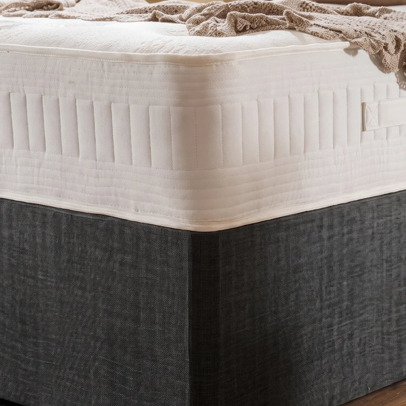 A close-up of a mattress with a white quilted top and a dark charcoal gray fabric-covered base, shown with a beige throw blanket draped across the corner.