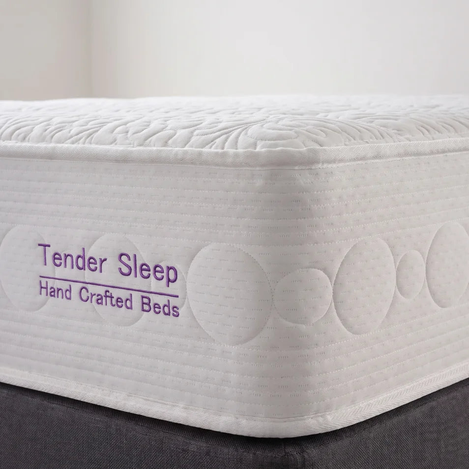 A close-up of a white mattress corner with quilted circular and leaf-like stitching, showing the text 'Tender Sleep Hand Crafted Beds' in purple on the side. The mattress sits on a dark gray bed base against a light neutral background.