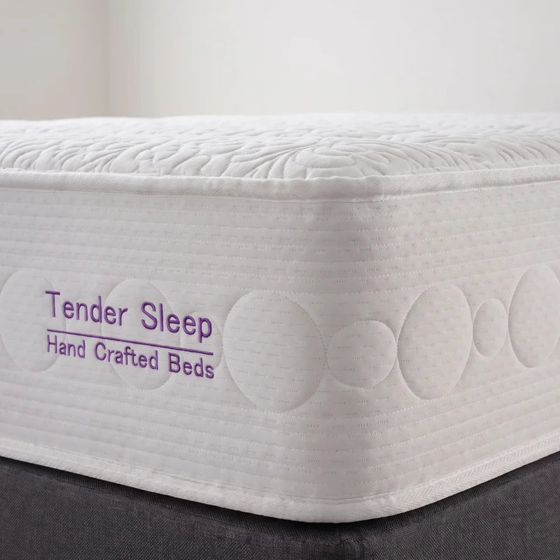 A close-up of a white mattress corner with quilted circular and leaf-like stitching, showing the text 'Tender Sleep Hand Crafted Beds' in purple on the side. The mattress sits on a dark gray bed base against a light neutral background.