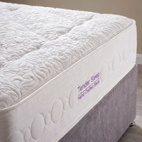 A close-up of a white quilted mattress topper or mattress edge with tufted wave patterns, attached to a gray upholstered bed base. Purple text on the side reads 'Tender Sleep Hand Crafted Beds'.