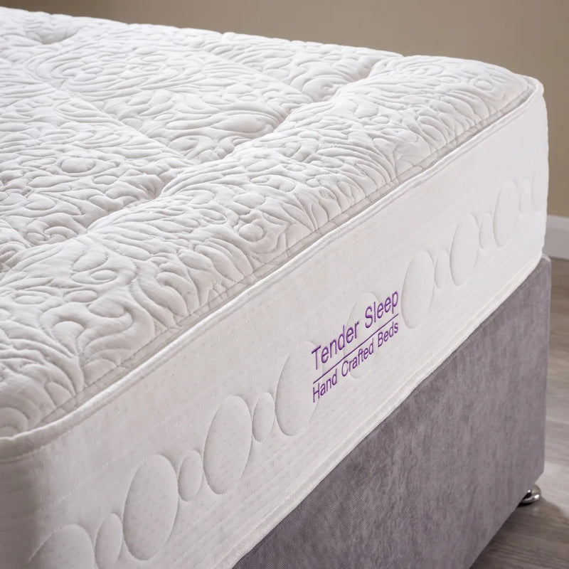 A close-up of a white quilted mattress topper or mattress edge with tufted wave patterns, attached to a gray upholstered bed base. Purple text on the side reads 'Tender Sleep Hand Crafted Beds'.