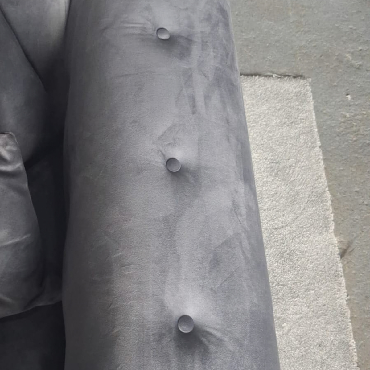Close-up of a gray upholstered armrest with button tufting, photographed against a concrete floor and adjacent furniture.