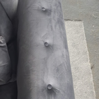 Close-up of a gray upholstered armrest with button tufting, photographed against a concrete floor and adjacent furniture.