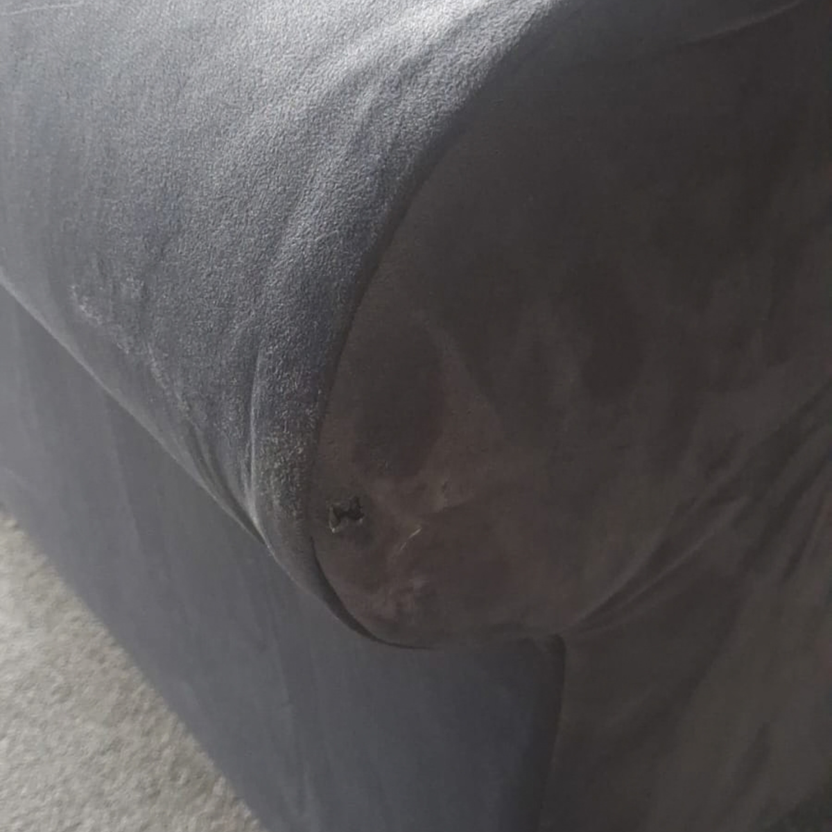 Close-up of a dark gray upholstered armrest or sofa corner with visible wear, stains, and a small dark spot on the fabric.