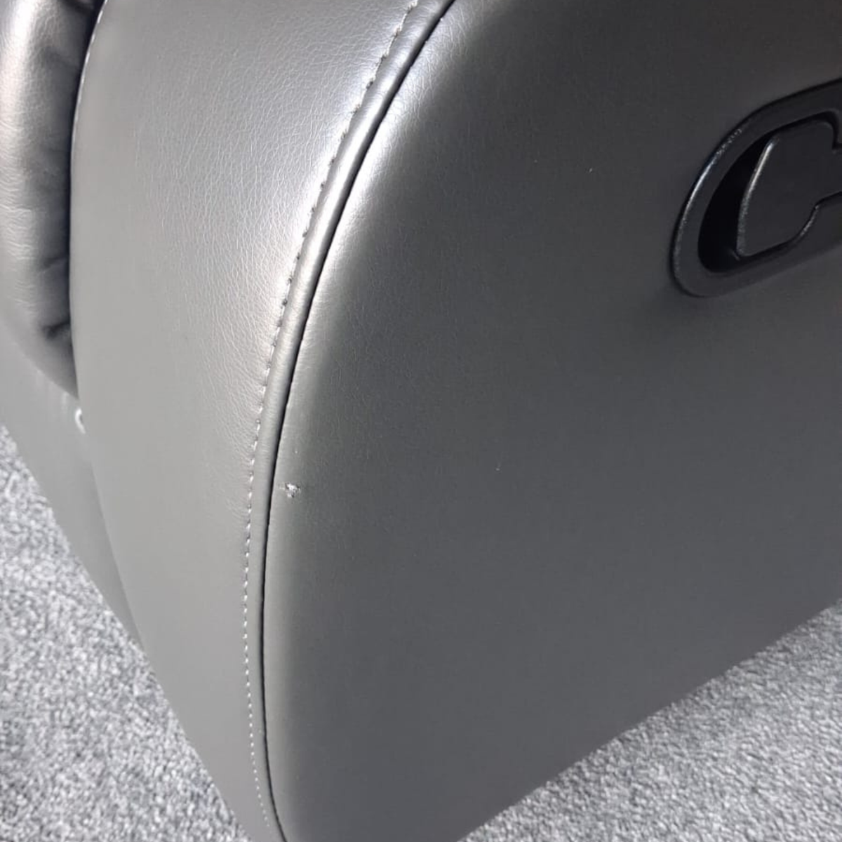 Close-up of a dark gray upholstered chair or recliner seat with visible stitching and a black control panel on the side, set on a gray carpeted floor.