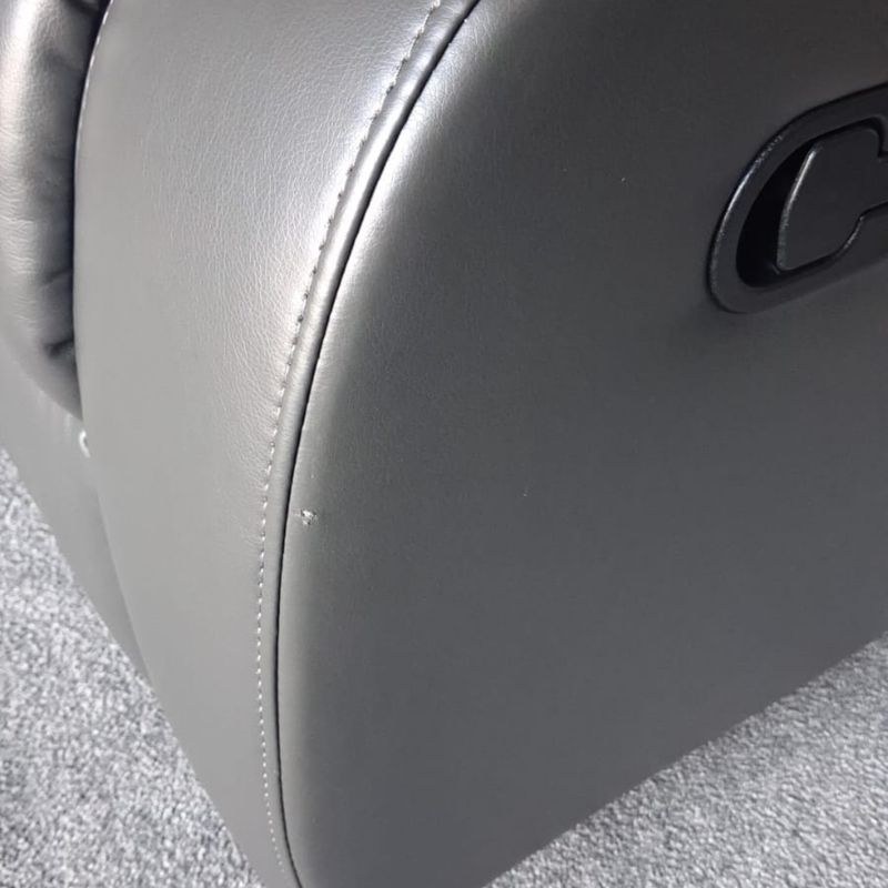 Close-up of a dark gray upholstered chair or recliner seat with visible stitching and a black control panel on the side, set on a gray carpeted floor.