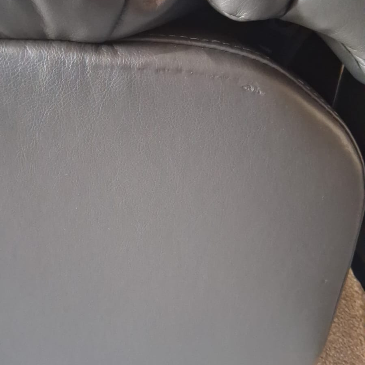 Close-up of a gray upholstered chair or cushion with visible leather or faux-leather texture, rounded edges, and a dark background.