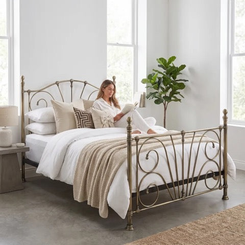 A woman in a white robe sits on a bed with white linens and a beige throw blanket, reading a book. The bed has an antique brass metal frame with decorative scrollwork, set in a bright bedroom with large windows, a side table, and a potted plant.