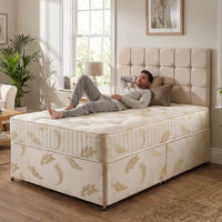 A man relaxes on a tufted divan bed with a cream upholstered headboard and base, decorated with a light beige leaf pattern. The scene is set in a bright, modern bedroom with a window, lamp, and houseplant.