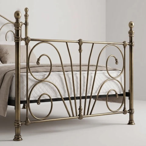 An ornate metal bed frame in a warm antique brass finish, shown with white bedding in a minimalist bedroom setting.