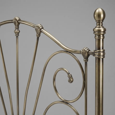 Close-up of an ornate metal bed frame headboard in a warm brushed brass or bronze finish, showing curved scrollwork and a vertical post against a plain gray background.