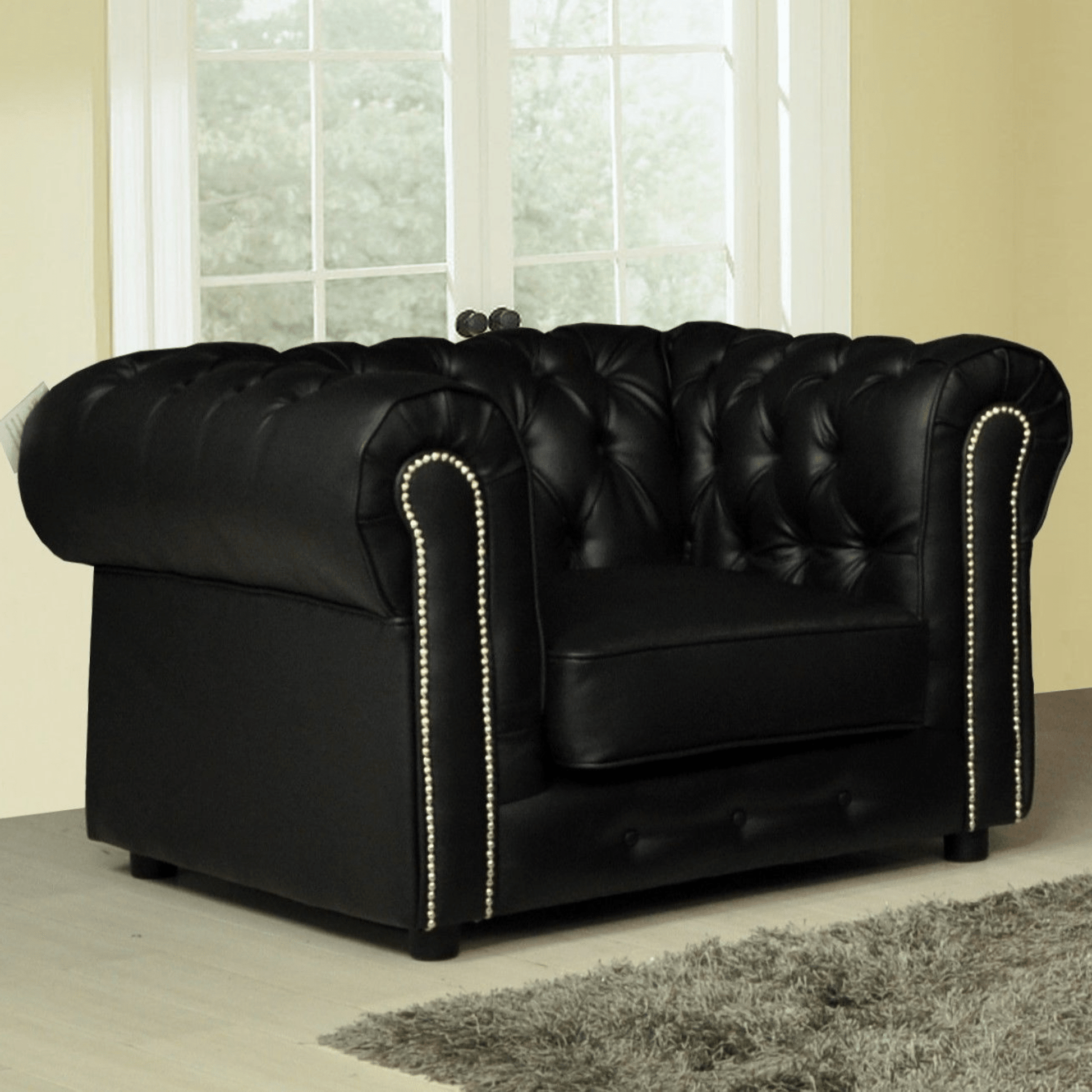 Black leather sofa with tufted backrest and nailhead trim in a room with large windows.