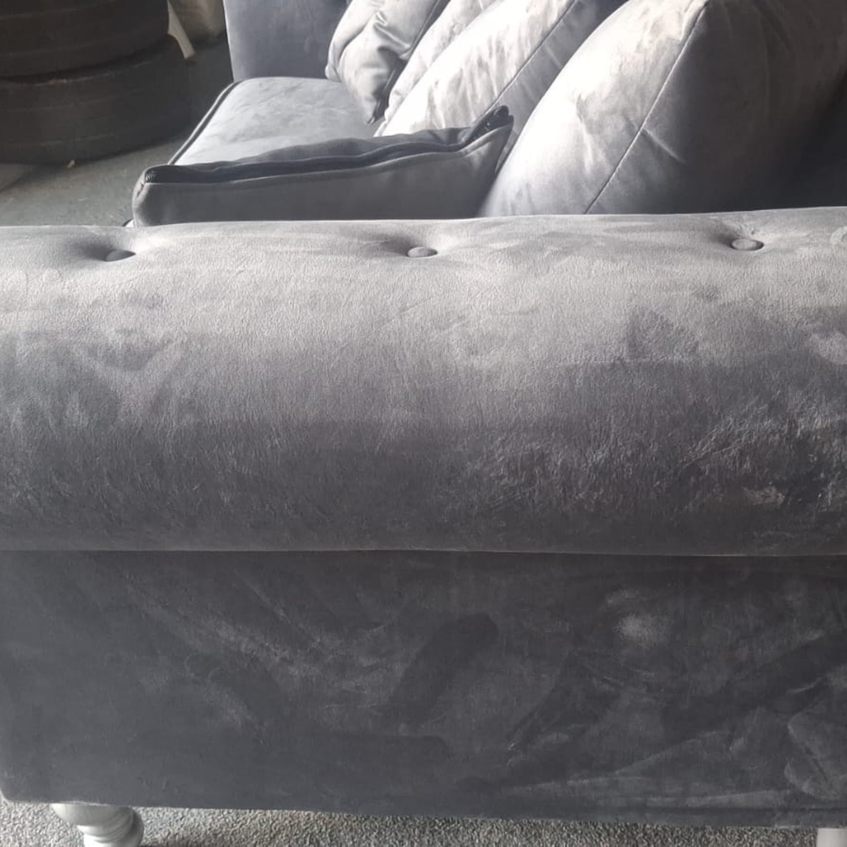 Close-up of a dark gray velvet or microfiber upholstered sofa section, showing the tufted armrest and side panel with visible wear marks and scuffs.