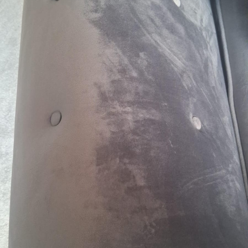 A close-up of a dark gray upholstered surface with visible wear, scuffs, and a small circular hole, photographed against a light gray floor/background.